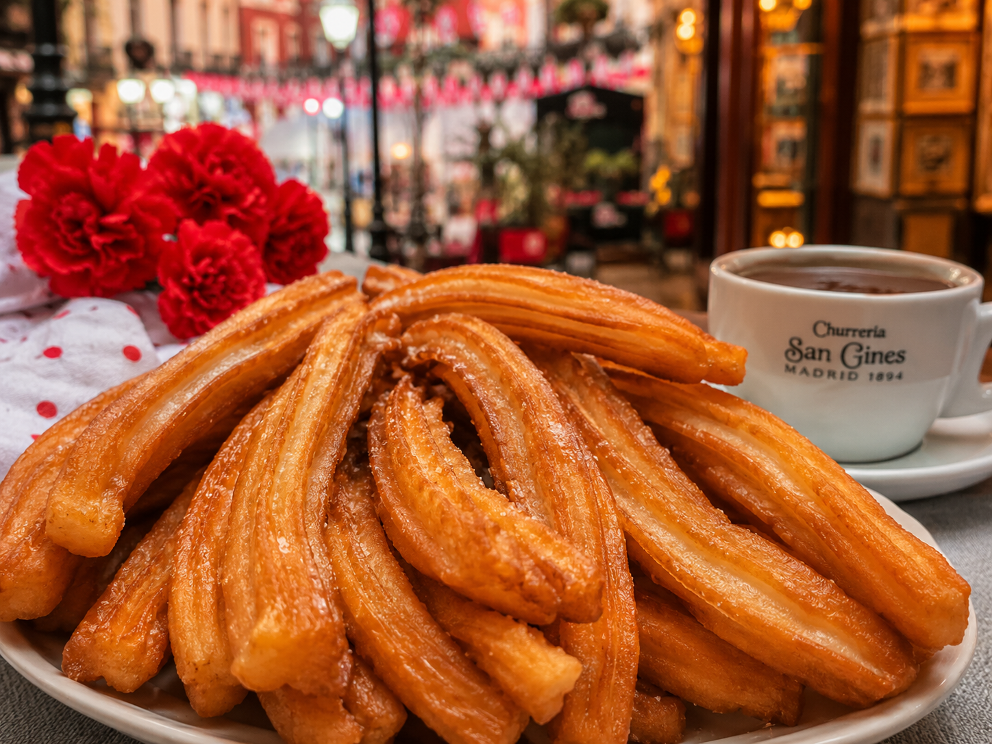 Churros recién hechos con textura crujiente, fondo con claveles rojos y ambiente festivo